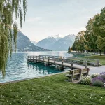 Vue panoramique du Lac d'Annecy depuis un parc public avec passerelle en bois et montagnes enneigées en arrière-plan